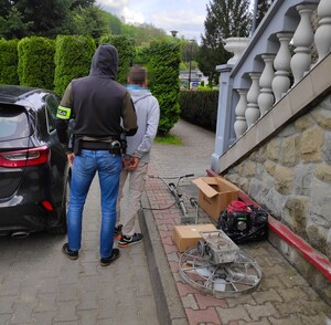 nieumundurowany policjant prowadzi zatrzymanego obok kilka odzyskanych przedmiotów pochodzących z włamania