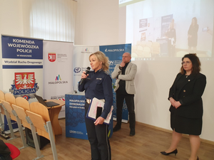 Policjantka trzymając w ręku mikrofon odpowiada na pytania. Za policjantką stoi kobieta i męźczyzna