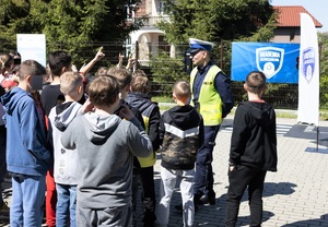 Umundurowany policjant ruchu drogowego w kamizelce odblaskowej z napisem Policja. Przed nim grupa dzieci, część z nich podnosi rękę do góry.
