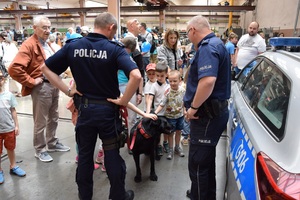 2 dwaj policjanci z psem służbowym stoją przy radiowozie, obok stoją dzieci i dorośli