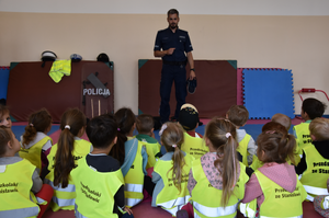 Umundurowany policjant oraz przedszkolaki siedzące na macie. Przedszkolaki mają na sobie kamizelki odblaskowe