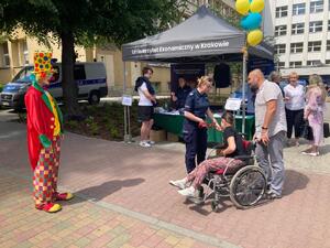 policjantka stoi obok mężczyzny i osoby na wózku inwalidzkim. Po lewej stronie osoba przebrana za klauna, w tle policyjne stoisko profilaktyczne