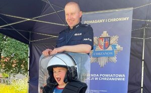 umundurowany policjant stoi z dzieckiem ubranym w kask i trzymajacy pałkę, z tyłu stoi rolap z napisem pomagamy i chronimy, na ziemi porozkładane sa pachołki