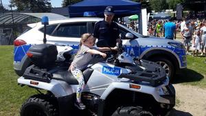 Dziewczynka siedzi na quadzie policyjnym. Obok stoi policjant w tle radiowóz policyjny