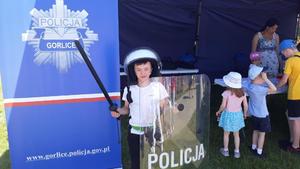Przy stoisku policyjnum i logo z napisem Policja Gorlice stoi chłopiec w kasku trzymający wyposażenie policyjne.