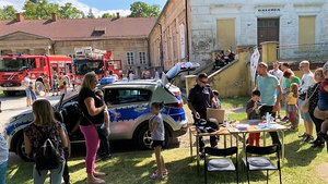 Na zdjęciu widać policyjny radiowóz i policjanta stoliki na których są poukładane ulotki i sprzęt policyjny wokoło tłum osób. W tle samochody