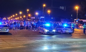 policjanci stojący obok radiowozó z włączonymi sygnałami świetlnymi. Za nimi widać tłum osób. Zdjęcie zrobione w porze nocnej