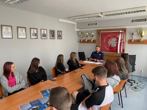 policjant siedzący za biurkiem, za min sztandar policji. Przed mundurowym uczniowie siedzący przy stole, a na nim broszuty