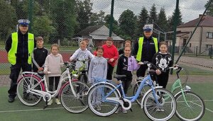 umundurowani policjanci ruchu drogowego na boisku szkolnym z dziećmi biorącymi udział w egzaminie, przed dziećmi 3 rowery w jasnych kolorach