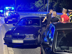 trzy samochody osobowe na poboczu drogi, obok których stoją osoby, z którymi rozmawiają policjanci. W tle widać radiowóz z włączonymi sygnałami świetlnymi