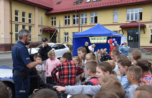 Umundurowany policjant z policyjną tarczą oraz ucznioiwe biorący udział w spotkaniu profilaktycznym