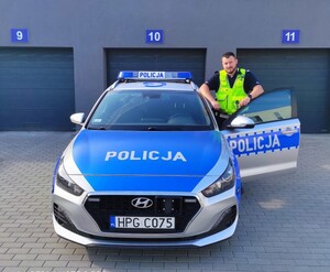 Na placu komendy, przed garażami stoi umundurowany policjant ruchu drogowego obok oznakowanego radiowozu. Policjant spiera się na otwartych drzwiach pojazdu od strony kierowcy