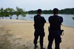 dwóch policjantów stojących tyłem na plaży, przed nimi zalew w którym pływają kajaki