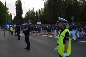 Policjantka wraz z policjantami zabezpieczają ulice w trakcie pochodu zawodników. W tle widać zawodników różnych państw.