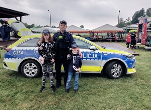 Na zdjęciu umundurowany policjant obok którego stoi po lewej stronie dziewczynka a po prawej chłopiec. Osoby stoja na tle oznakowanego radiowozu. W tle rozłożone namioty i siedzący ludzie oraz wóz strażacki.jpg