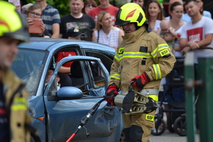 Strażak OSP podłączający hydrauliczne nożyce do cięcia, podczas pokazu ratownictwa. W tle samochód z poszkodowanym i ludzie obserwujący pokaz