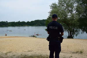 policjant stojący tyłem do zdjęcia, na plaży, przed nim zalew w którym pływają kajaki