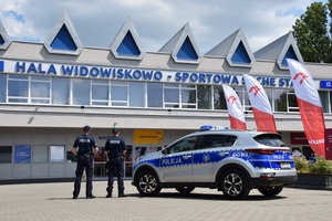 dwóch umundurowanych policjantów stojących obok radiowozu. Za nimi widać Halę Widowiskowo-Sportową ,,Suche Stawy''