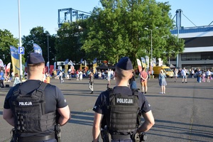 dwóch umundurowanych policjantów stojących tyłem do zdjęcia przy stadionie na ul. Reymonta. W oddali widać tłum ludzi