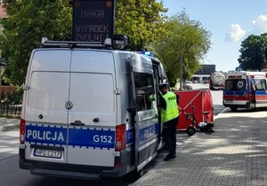 umundurowany policjant stojący przy radiwozie. Przed radiwozem widać rozłożony parawan oraz samochód pogotwia ratunkowego