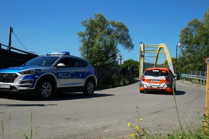 stojący oznakowany radiowóz i jadacy białopomarńczowy samochód rajdowy na metalowy w kierunku dużego mostu