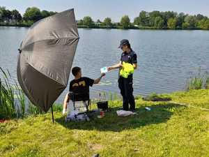 Policjantka podaje wędkarzowi ulotkę. Wędkarz siedzi na stołku przed nim wędki. Po lewej stronie wędkarza parasol. W tle zalew.