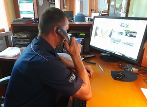 Policjant  siedzi przy biurku dyżurnego, przy uchu trzyma słuchawkę