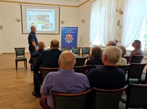 Naczelnik Wydziału Prewencji przedstawia zaganienie debaty, przed min uczestnicy oraz Komendanta powiatowy siedząna krzesłach, w tle prezentacja