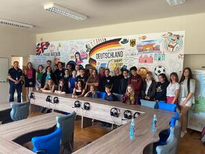 Młodziez polsko-niemiecka wypoczywajaca w Jaroszowcu wracz z policjantem i pracownikiem sanepidu. Zdjęcie w sali. w tle kolorowa plansza z flagą niemiecką i rózne malunki z atrakcyjnymi miejscami.