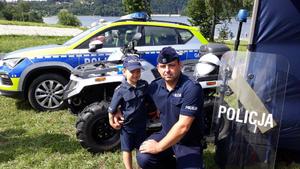 Policjant wraz z dzieckiem ubranym w strój policjanta. Za nimi stoi quad i radiowóz policyjny. Obok stoi tarcza policyjna. W tle jezioro.