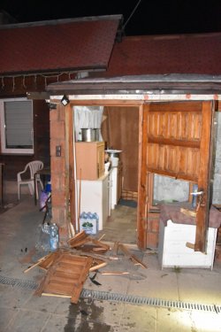 Uszkodzone drewniane drzwi znajdujące się przy  wejściu do budynku (ganku). Część z nich znajduje się na kostce brukowej. Za drzwiami widać meble na których znajdują się m.in. garnki.