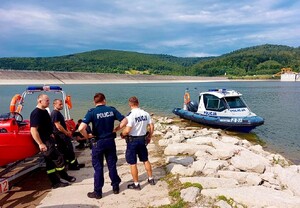 Po prawej stronie zdjęcia, na wodzie jest łódź policyjna, natomiast po lewej łódź straży pożarnej obok której stoi dwóch strażaków i dwóch umundurowanych policjantów.