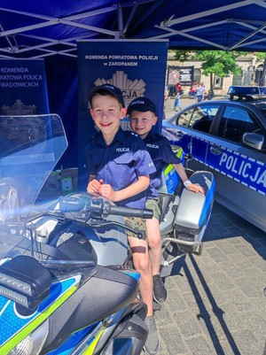 Dwóch małych chłopców ubranych w strój policjnata, siedza na motocyklu policyjnym. W tle policyjne banery oraz fragment radiowozu.