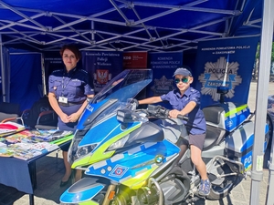Po lewej stronie umundurowana policjantka stoi obok policyjnego motocykla na którym siedzi chłopiec ubrany w strój policjanta. W tle policyjne banery.