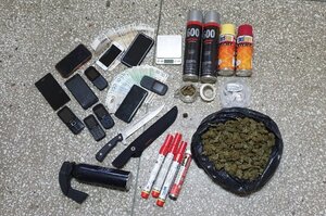 Ujawnione i zabezpieczone narkotyki i rzeczy leżące obok siebie. Susz roślinny, nóz, gaz pieprzowy, pieniądze, telefony komórkwe, spreje, race oraz waga elektryczna.