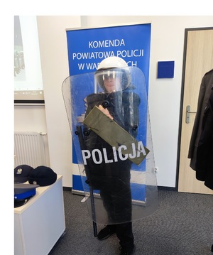 Chłopiec ubrany w kamizelkę kuloodporną, kask, w ręku trzyma pałkę oraz policyjną tarczę. Za nim baner policyjny z napisem Komenda Powiatowa Policji w Wadowicach