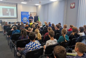 Dzieci siedzące na krzesłach tyłem w pomieszczeniu. Przed nimi stoi umundurowany policjant. W tle baner policyjny oraz elementy umundurowania i projektor na którym jest wyślwietlana prezentacja