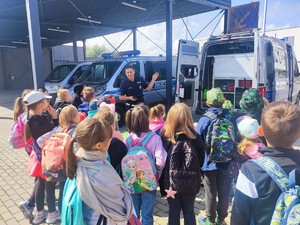 Dzieci stojace tyłem, przed nimi umundurowany policjant, który prezentuje oznakowany radiowóz. W tle dwa oznakowane radiowozy.