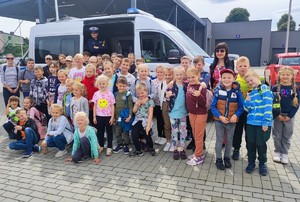 Grupa dzieci stojąca przed oznakowanym radiowozem w którym stoi umundurowany policjant. W tle budynek i zaparkowane samochody.