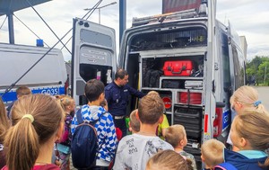 Grupa dzieci stojąca tyłem, przed nimi umundurowany policjant który pokazuje oznakowany radiowóz i sprzęt będący na jego wyposażeniu