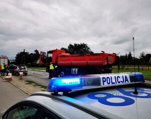 Miejsce zdarzenia drogowego - na pierwszym planie dach radiowozu z lampą błyskową z napisem Policja. W tle samochód ciężarowy i osobowy oraz  umundurowani policjanci