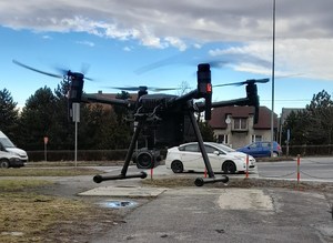 Dron unoszący się w powietrzu. W tle dron i poruszające się pojazdy oraz zabudowania