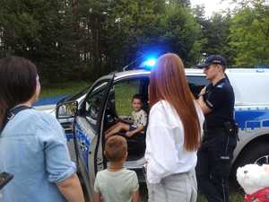 Dziecko za kierownica radiowozu policyjnego. Obok przy drzwiach stoi policjant i grupa dorosłych