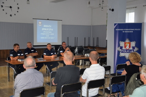 uczestnicy debaty siedzący tyłem do zdjęcia, przed nimi przedstawiciele Policji, za nimi wyświetlona prezentacja, obok po prawej baner KPP w Krakowie