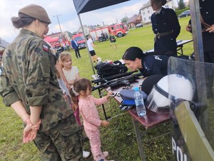 . Rajsko piknik historyczno - wojskowy policjantka prezentuje sprzęt na stoisku dziecko  ogląda kajdanki obok stoi kobieta w wojskowym mundurze
