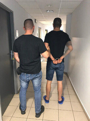 policjant kryminalny prowadzi osobę zatrzyamaną z założonymi kajdankami na ręce trzymane z tyłu przez korytarz policyjnej jednostki