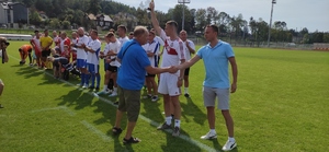 Prezes Ludowego Klubu Sportowego Kłos gratuluje piłkarzowi. W tle boisko i zawodnicy stojaccy w rzędzie