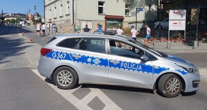 Na zdęciu widać oznakowany radiowóz zaparkowany na skrzyżownaiu w zwiąku z zbaezpieczeniem wadowickiego biegu. Za nim widać ucestników biegu. W t