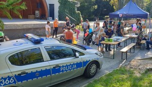 Po lewej stronie rozłozony namiot pod którym na stoliku poukładane są ulotki i sprzęt policyjny. Po prawej radiowóz i motocykl policyjny oraz umundurowany policjant i policjantka. Obok dwie osoby dorosłe i dzieci