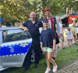 Dziewczynka w policyjnej czapce oraz mężczyzna w stroju krakowskim pozują do zdjęcia z policjantem przy radiowozie